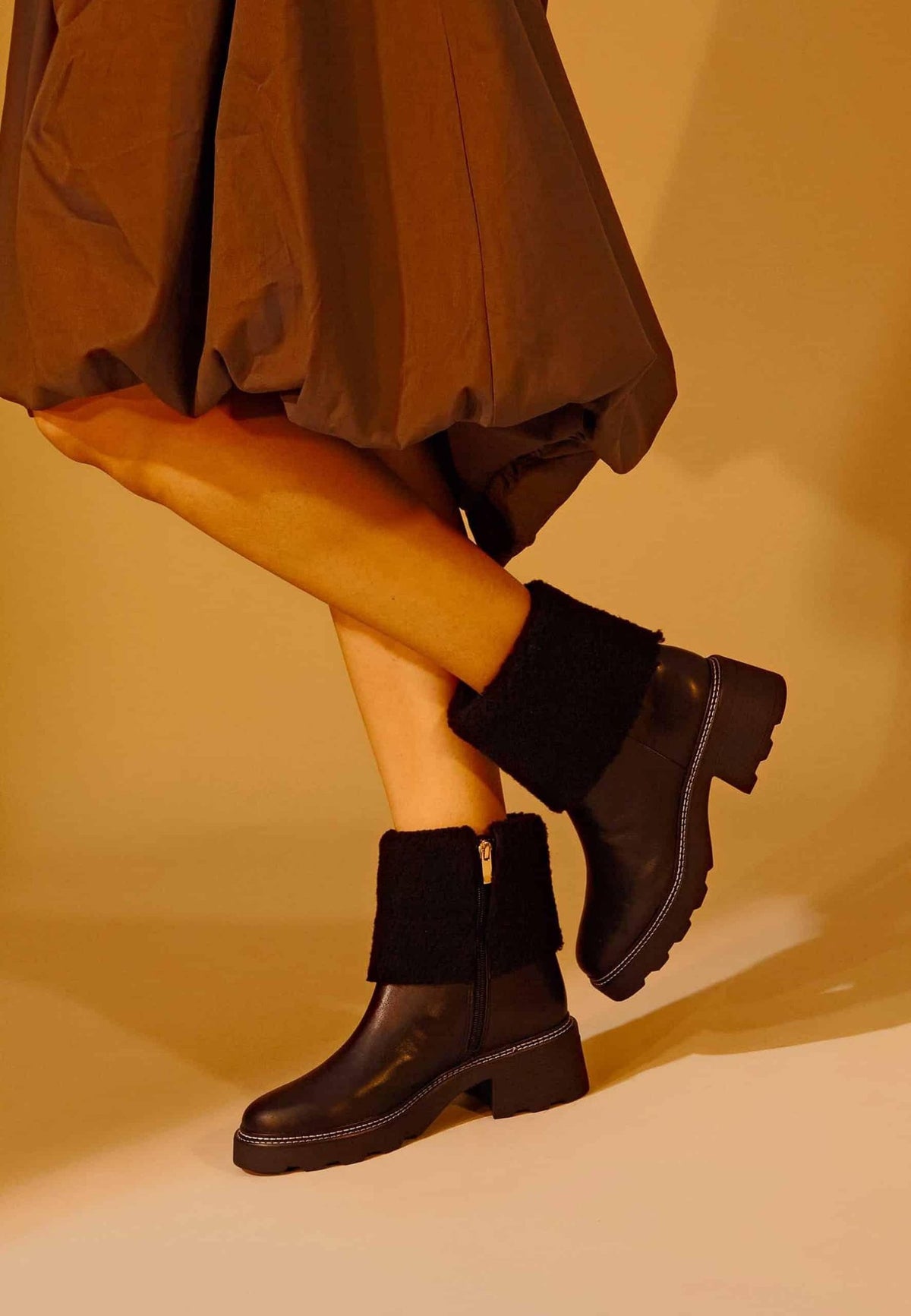 1. Woman wearing black leather ankle boots with shearling detail and block heel, styled with a brown skirt in a warm-toned setting