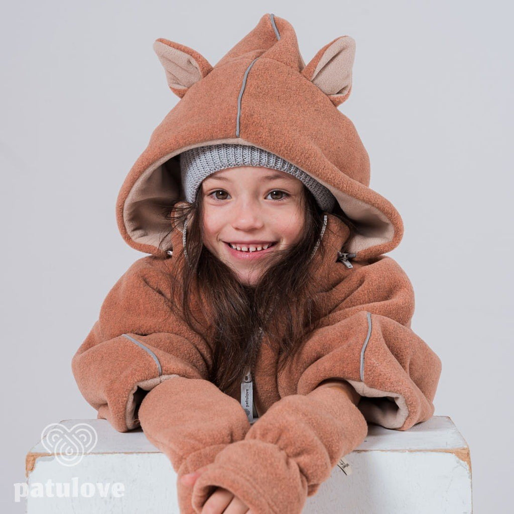 3. Child leaning on white box wearing Patulove fox car poncho with hood and grey beanie, smiling