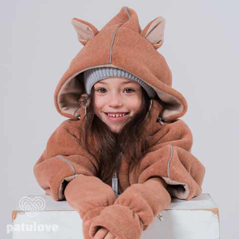 3. Child leaning on white box wearing Patulove fox car poncho with hood and grey beanie, smiling