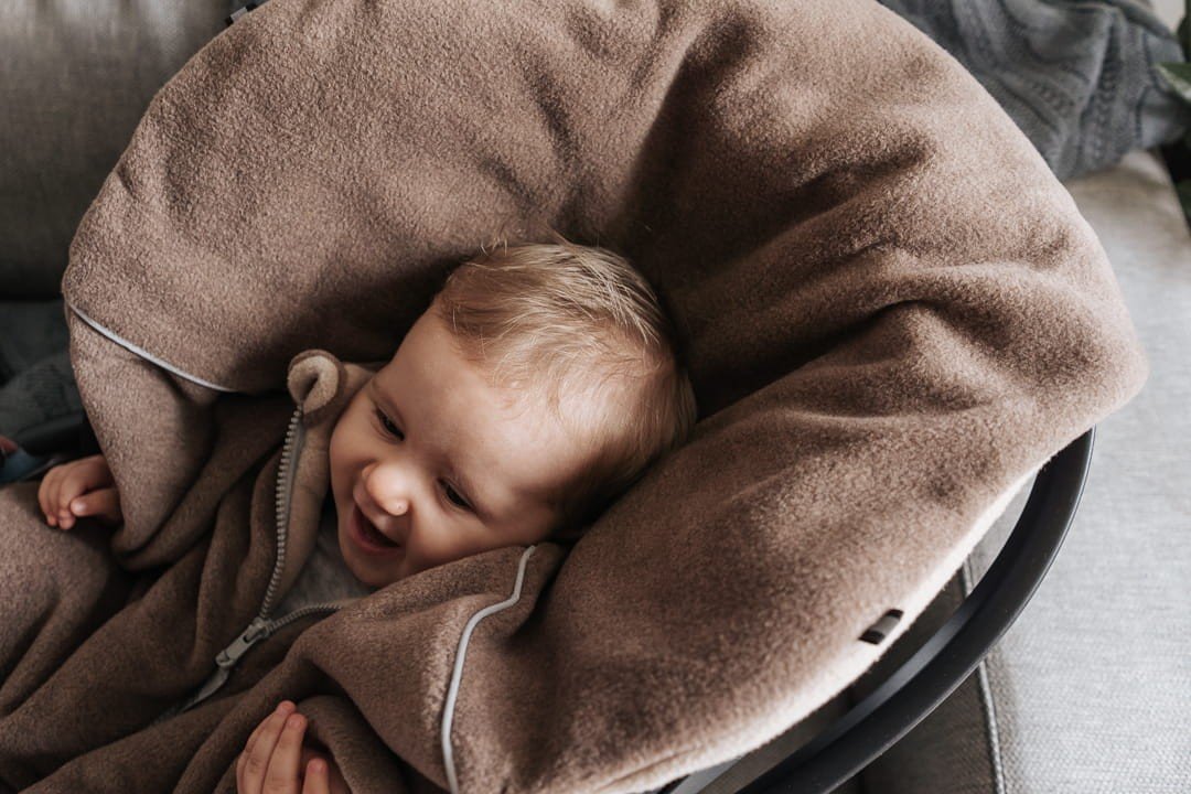 4. Baby smiling while wrapped in brown Patulove car poncho, lying on a soft surface