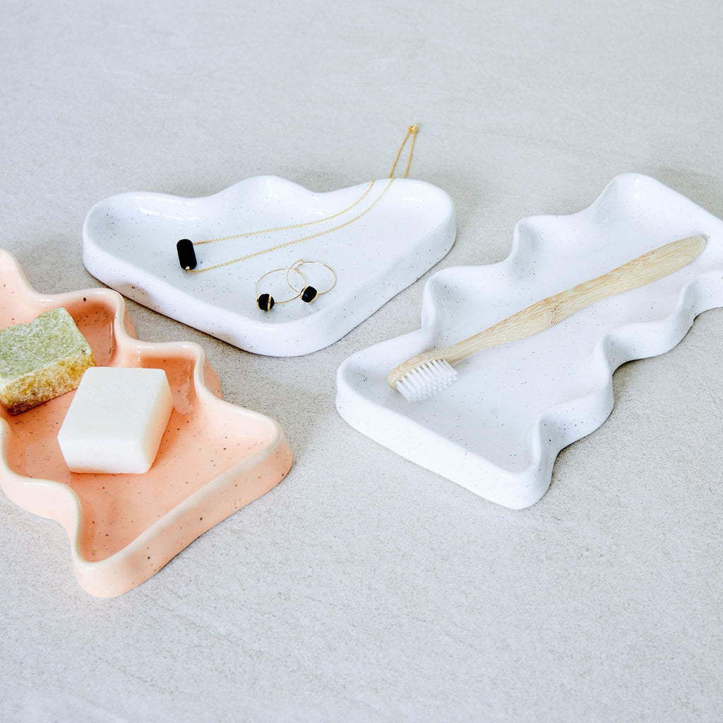 1. Lifestyle image of 5mm Paper Ceramic Wave Tray in white and peach, holding jewelry and toiletries on a bathroom counter
