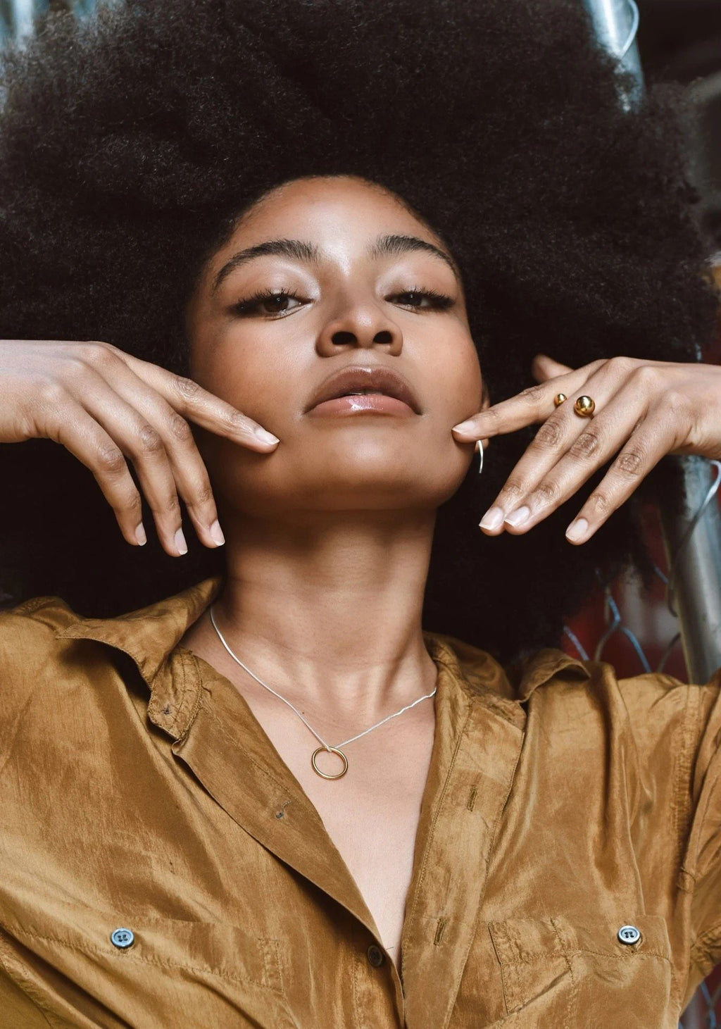 1. Woman wearing gold circle necklace with matching ring, styled with a brown shirt, showcasing minimalist jewelry in an urban setting