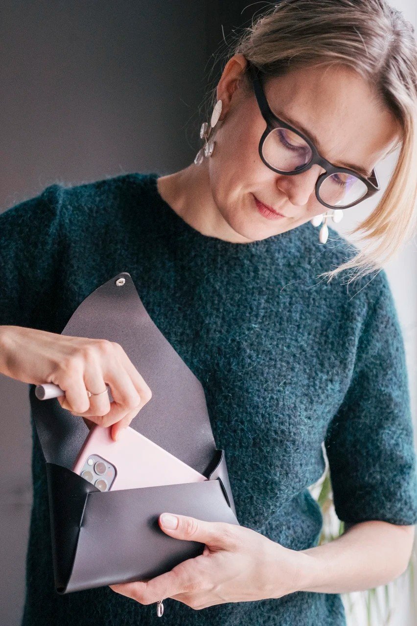 6. Woman placing smartphone into black leather clutch bag, wearing teal sweater, indoors