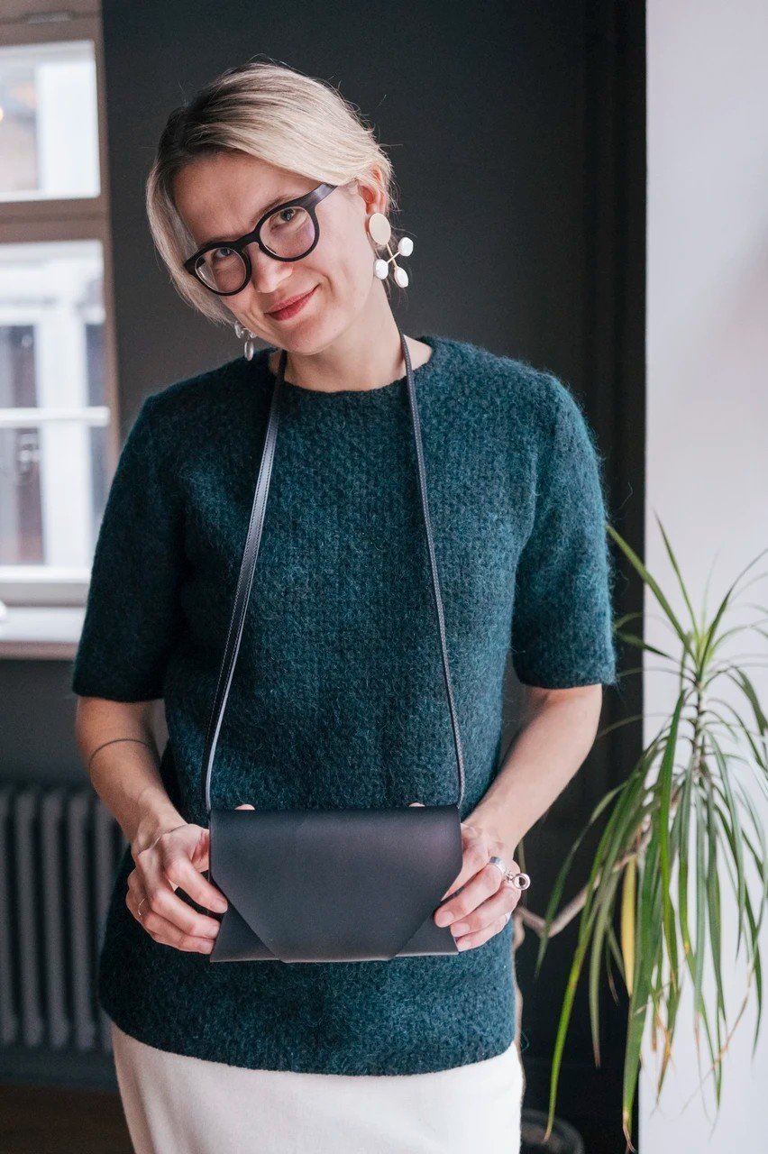 7. Woman wearing black leather clutch bag with long strap around neck, standing indoors