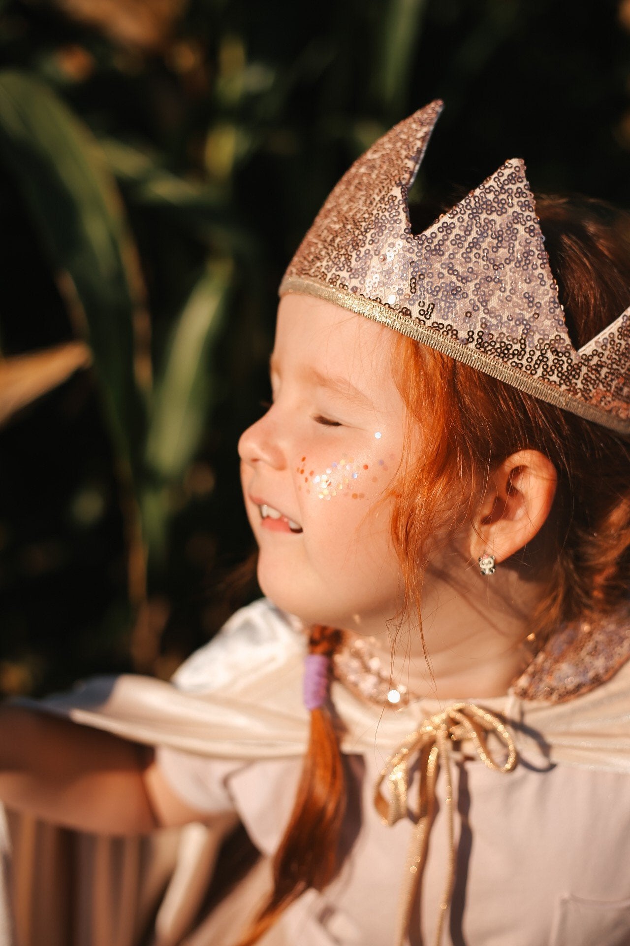 5. Young girl with red hair wearing a glittery crown and cape, smiling with gold star dust glitter on her cheek, in a natural setting