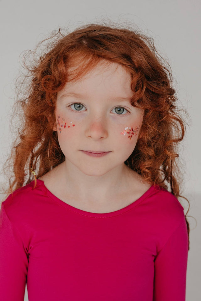 4. Young girl with red hair in pink top, front view, with moi mili Rainbow Star Dust glitter applied to her cheeks