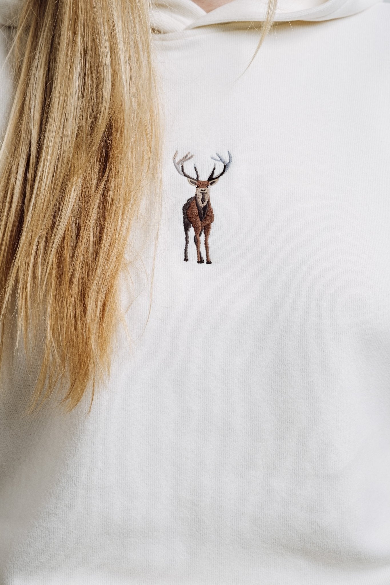 1. Close-up of cream Sossu Deer Hoodie with embroidered deer detail, worn by a woman
