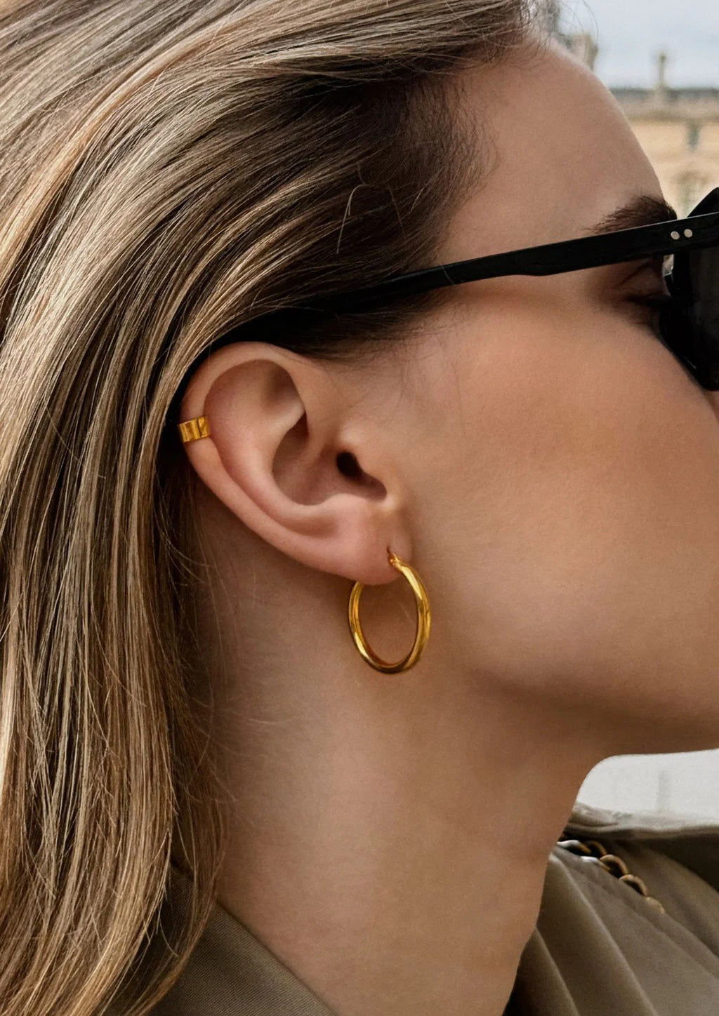 1. Woman wearing gold Django hoop earring by NO MORE, styled with sunglasses, showcasing elegant design and 24k gold vermeil finish
