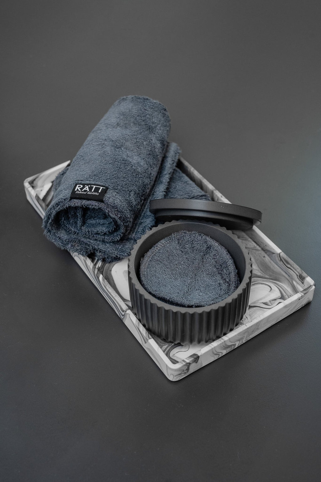 2. RÄTT Home Spa Set featuring black jesmonite container, dark gray bamboo terry towels, and cleansing pads on a marbled tray, shown on a dark background