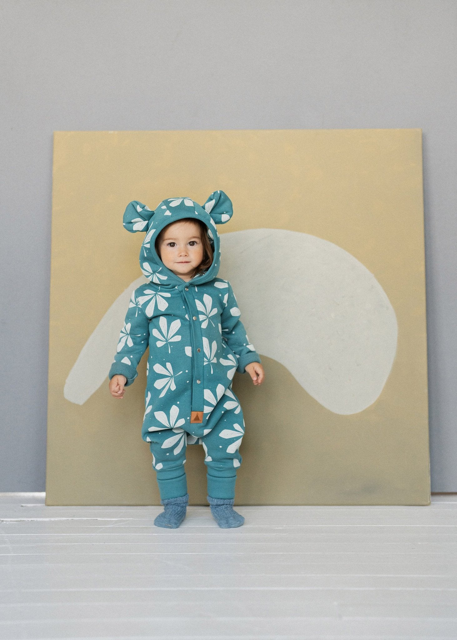 4. Child standing against beige backdrop in green jumpsuit with chestnut leaf print and hood with ears