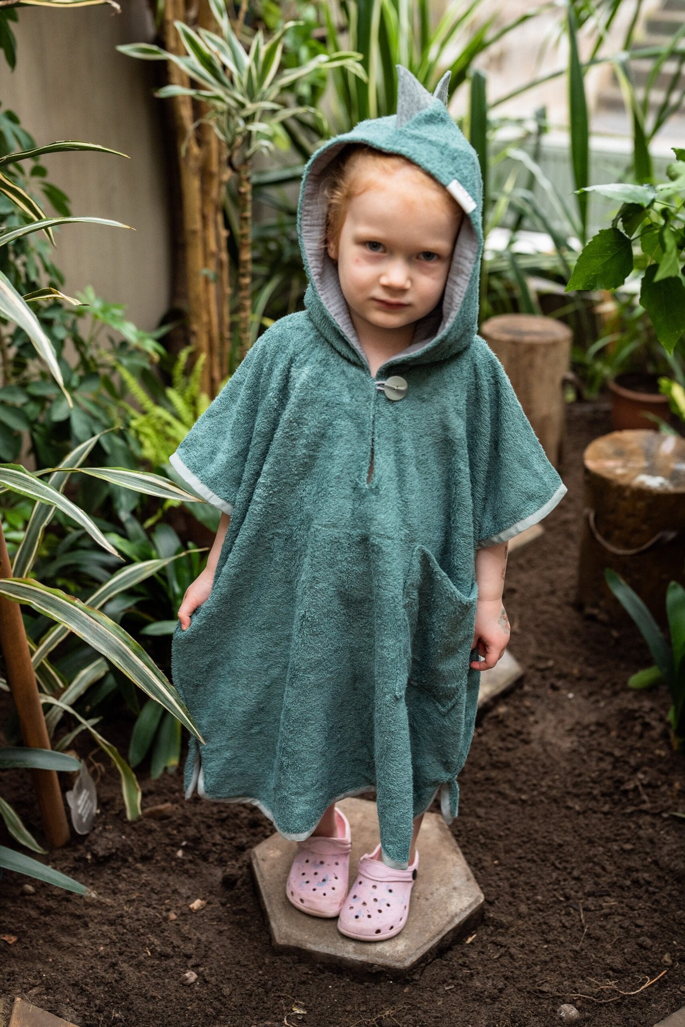 2. Toddler wearing green RÄTT short-sleeve dinosaur poncho with hood, standing in a garden