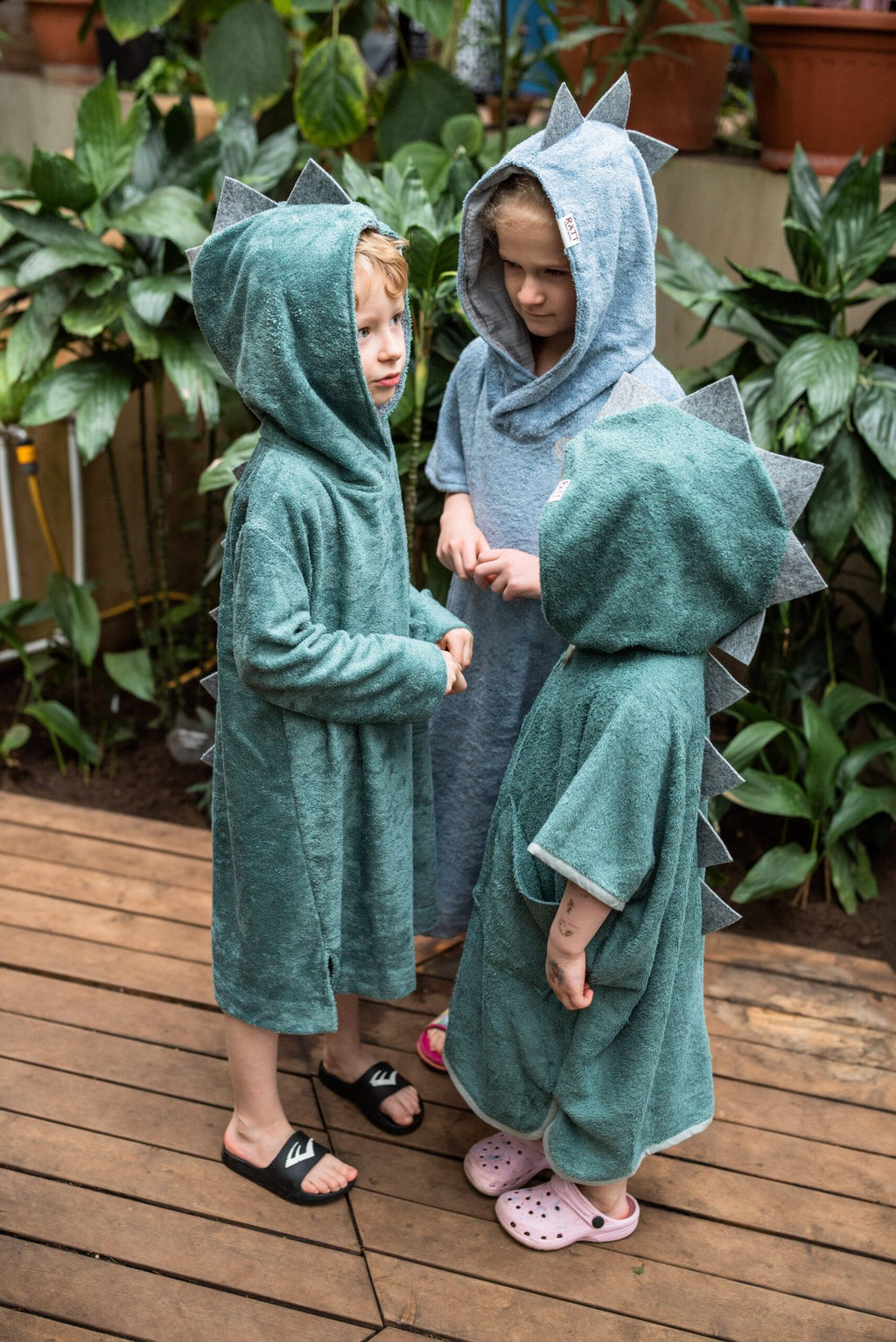 1. Kids wearing RÄTT short-sleeve dinosaur ponchos in green and blue, shown in a garden setting
