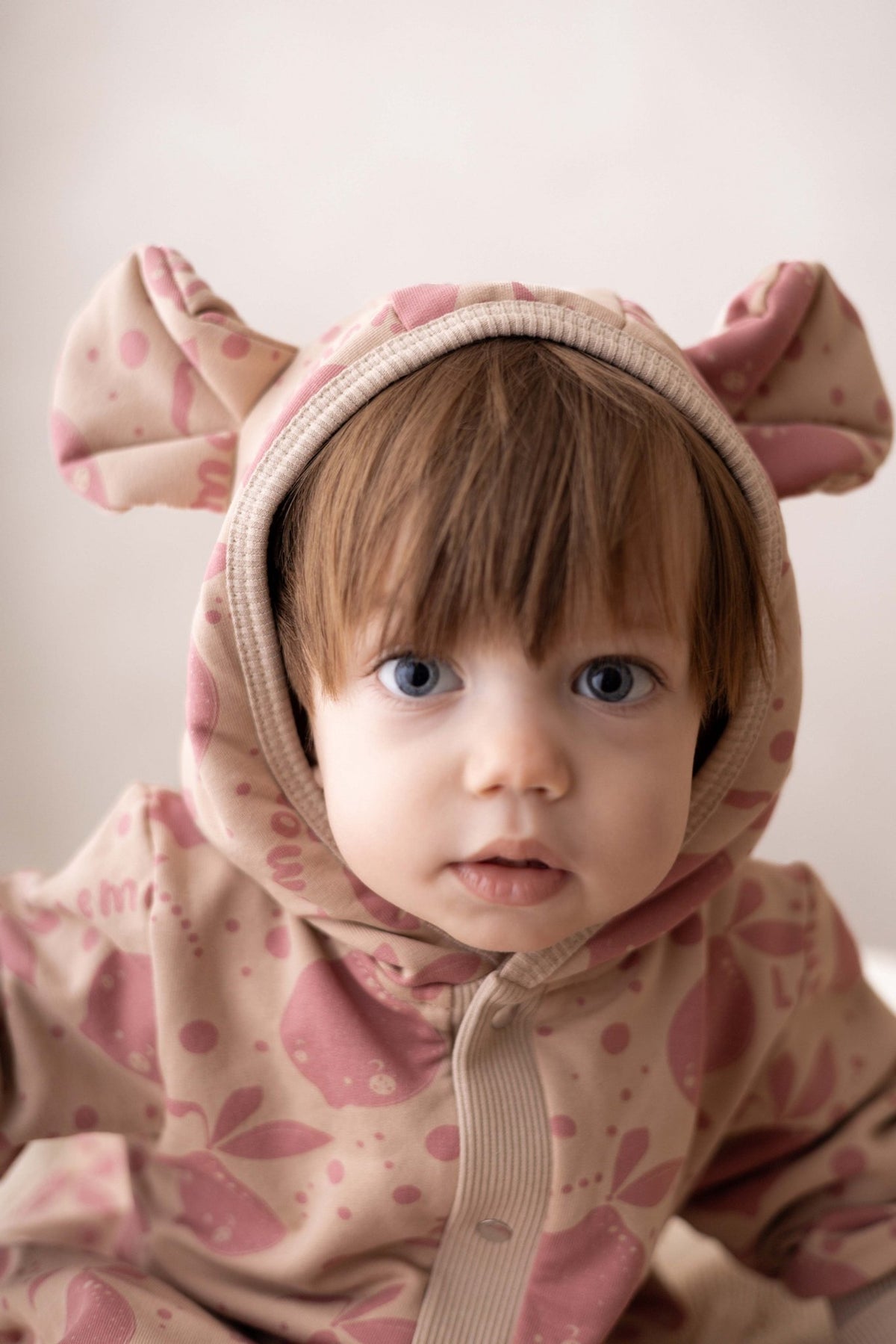 1. Baby wearing Zezuzulla beige jumpsuit with pink lemon print and hood with ears, sitting indoors