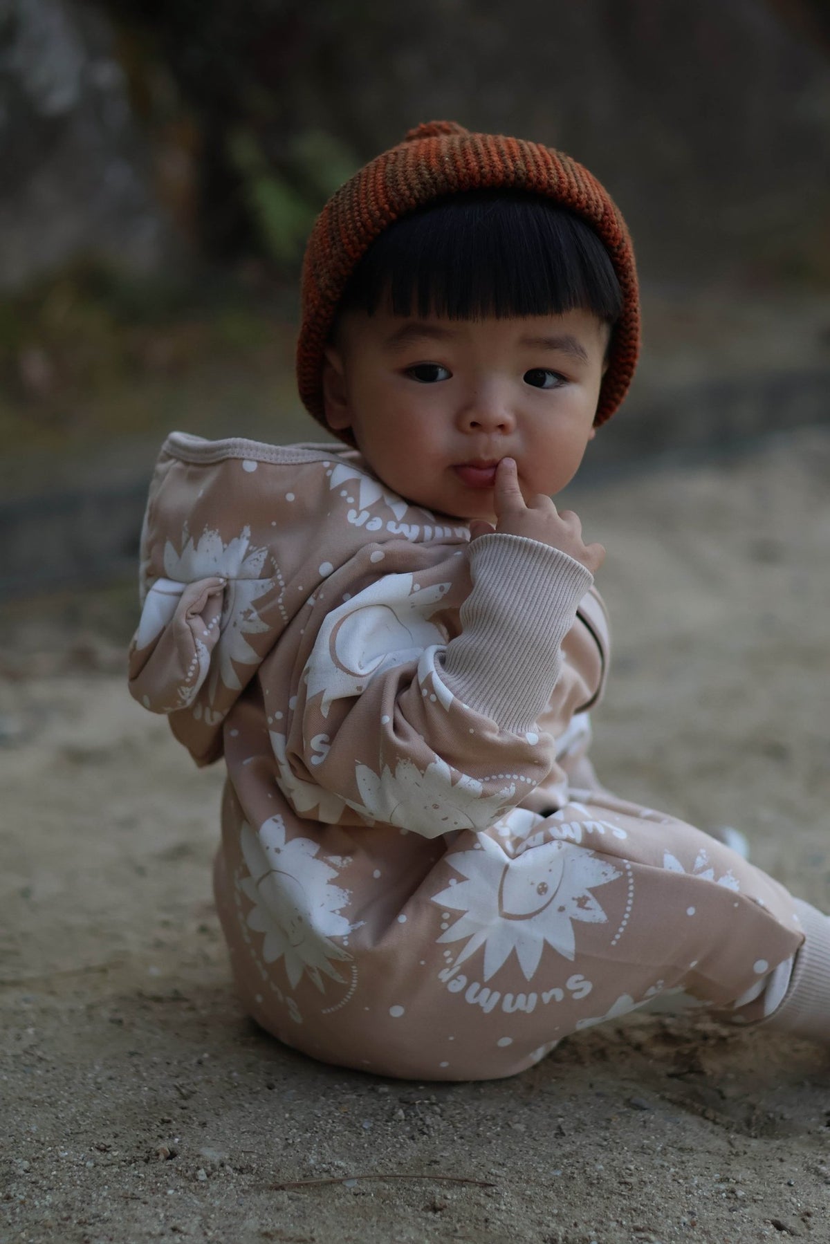 1. Baby sitting outdoors in beige sun-printed jumpsuit with hood and ears, wearing a brown beanie