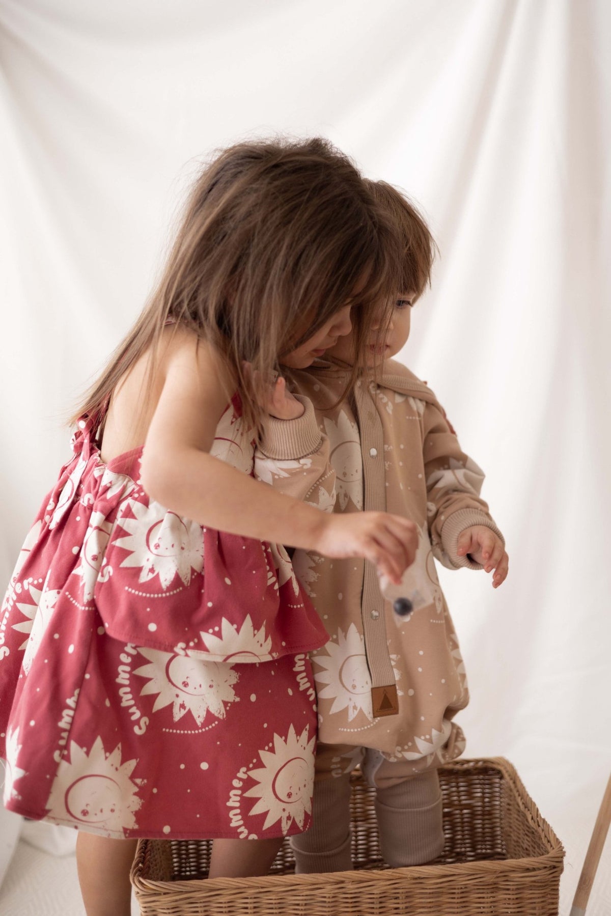 1. Two children in sun-printed outfits, one in beige jumpsuit with hood and ears, standing in basket