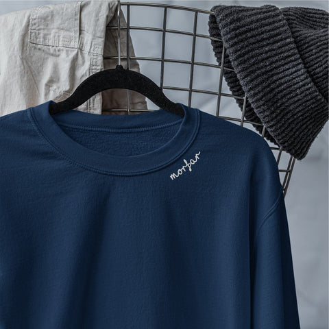 1. Navy sweatshirt with light gray 'Morfar' embroidery on collar hanging on a wire rack with folded clothes