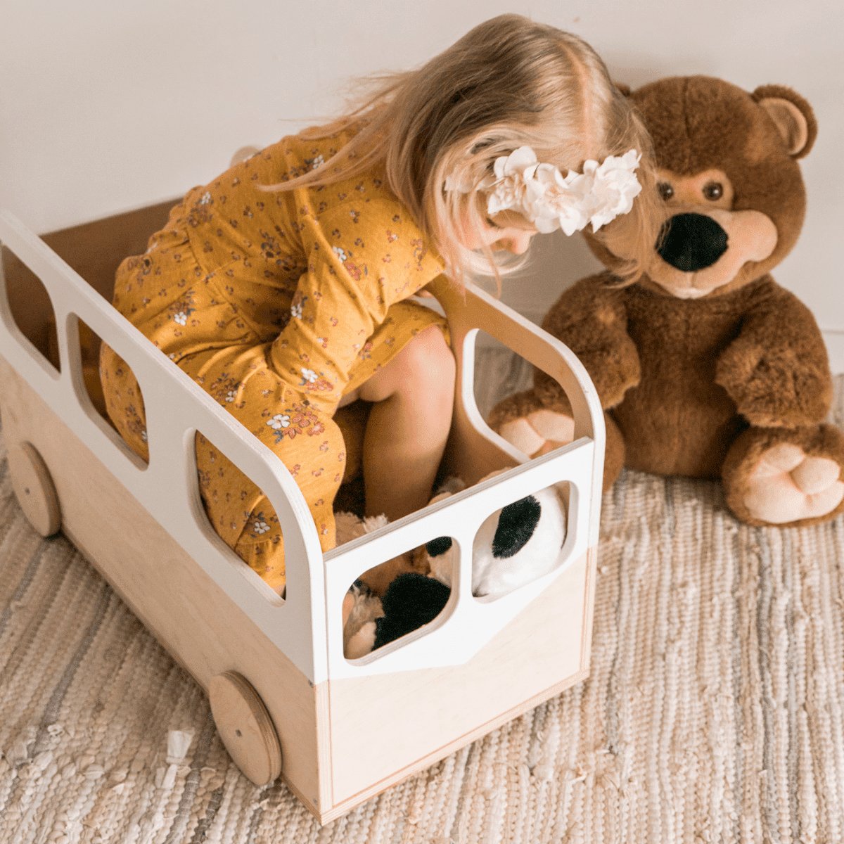 1. Child sitting inside Ewart Woods kids toy storage box on wheels, styled as a bus, with a teddy bear on a rug