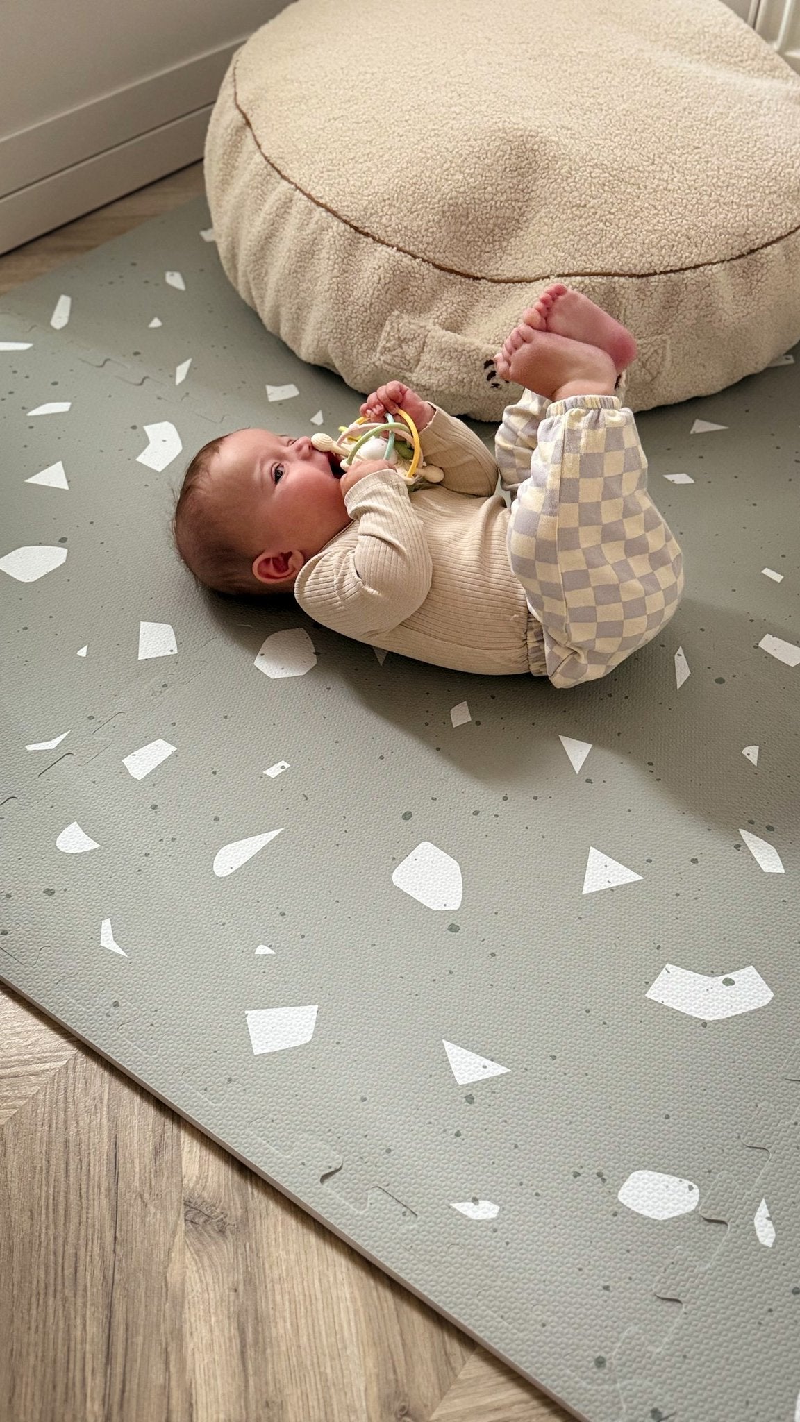 5. Baby on OLYA KIDS Terrazzo Sage playmat with beige cushion, wearing checkered pants, holding a toy.
