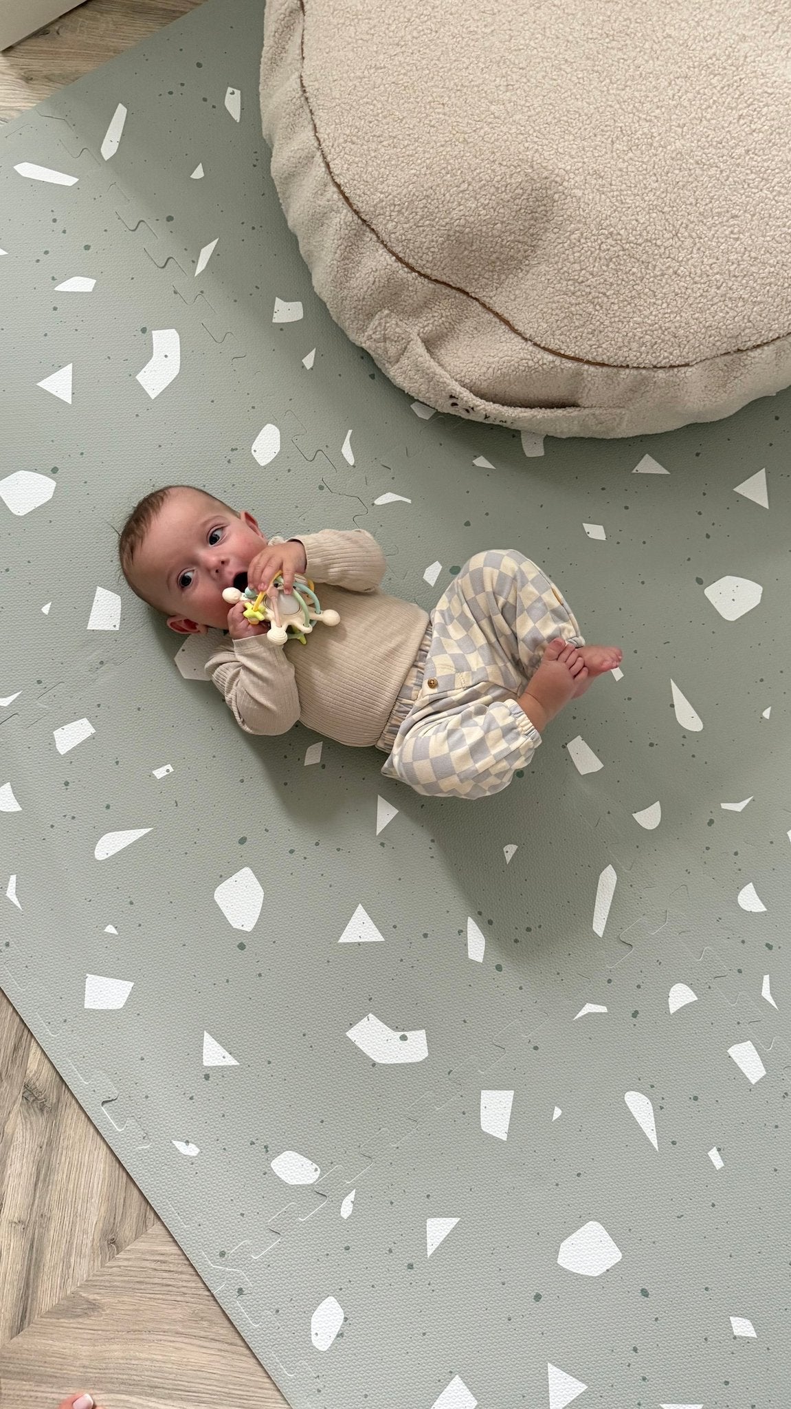 6. Baby on OLYA KIDS Terrazzo Sage playmat with beige cushion, wearing checkered pants, holding a toy.