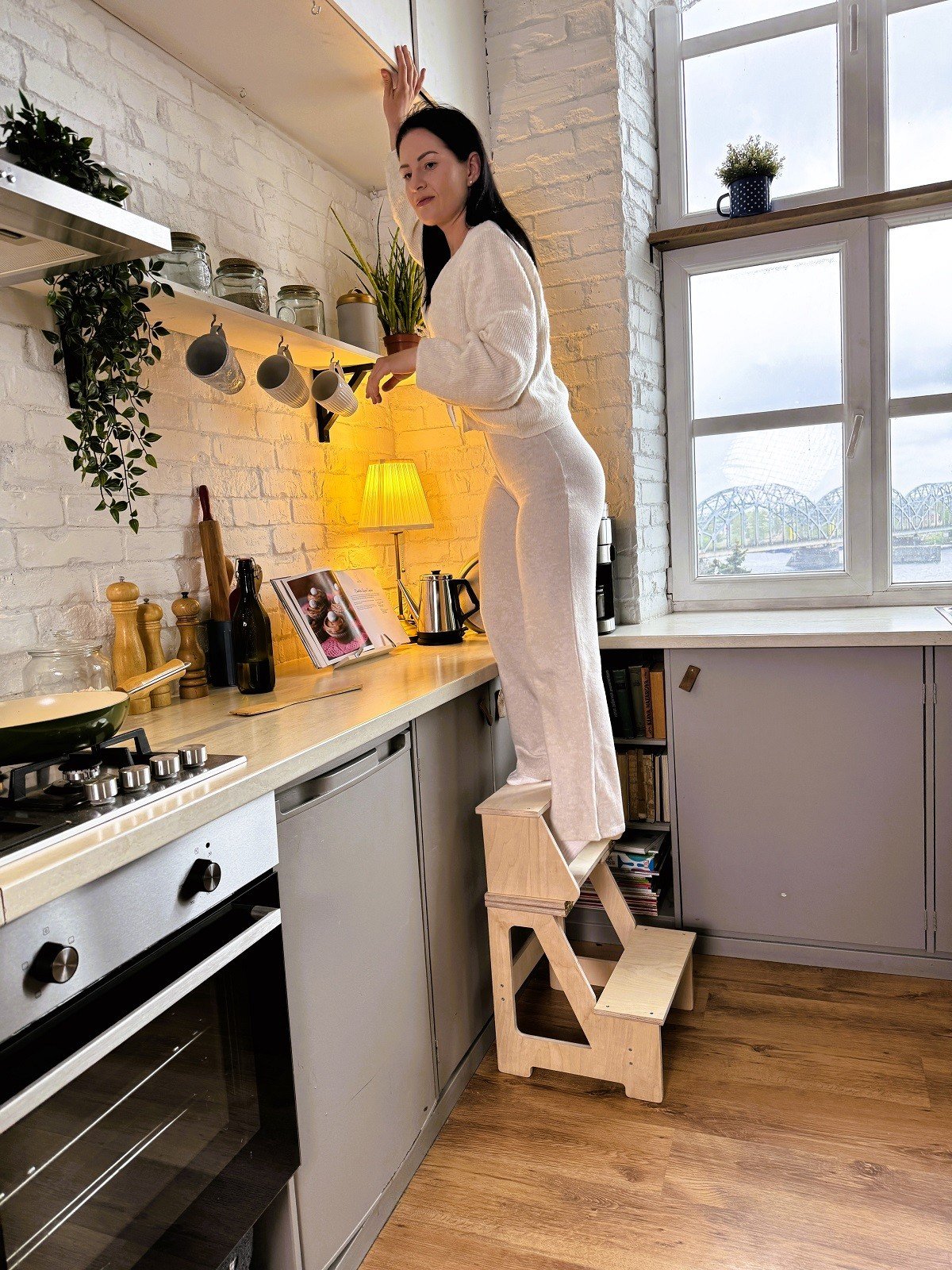 1. Woman standing on wooden folding seat and climbing helper in kitchen to reach shelf