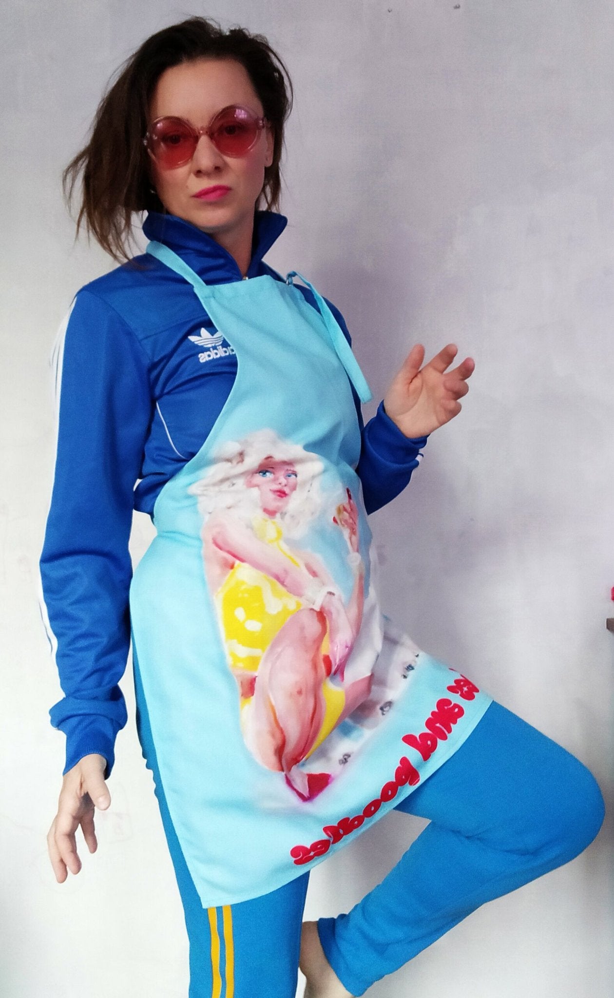 1. Woman posing in Katrin Valgemäe apron with blonde woman and poodle artwork, wearing blue tracksuit and pink sunglasses