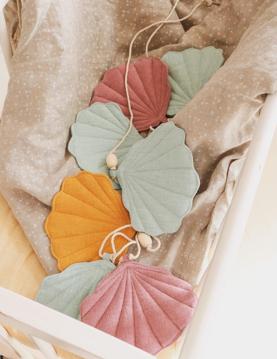 3. Lifestyle image of moi mili Shell Garland with mint, dirty pink, and mango linen shells draped over a patterned fabric in a cozy setting