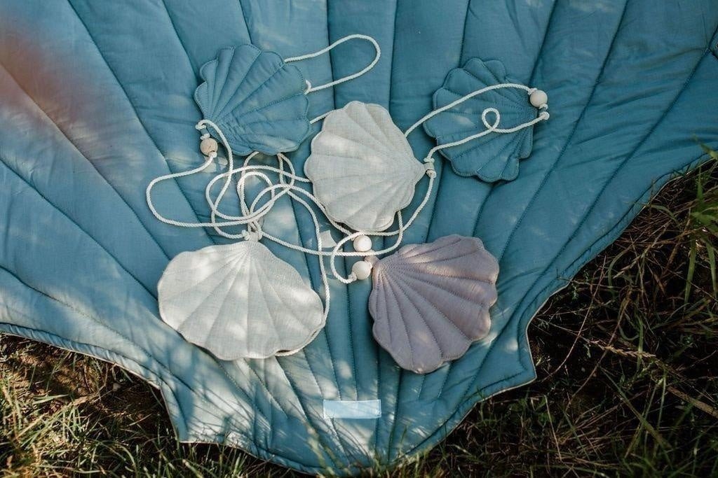 1. Shell garland with linen pendants in sky blue, grey, and desert sand on a cotton string with wooden beads, displayed on a blue quilt outdoors