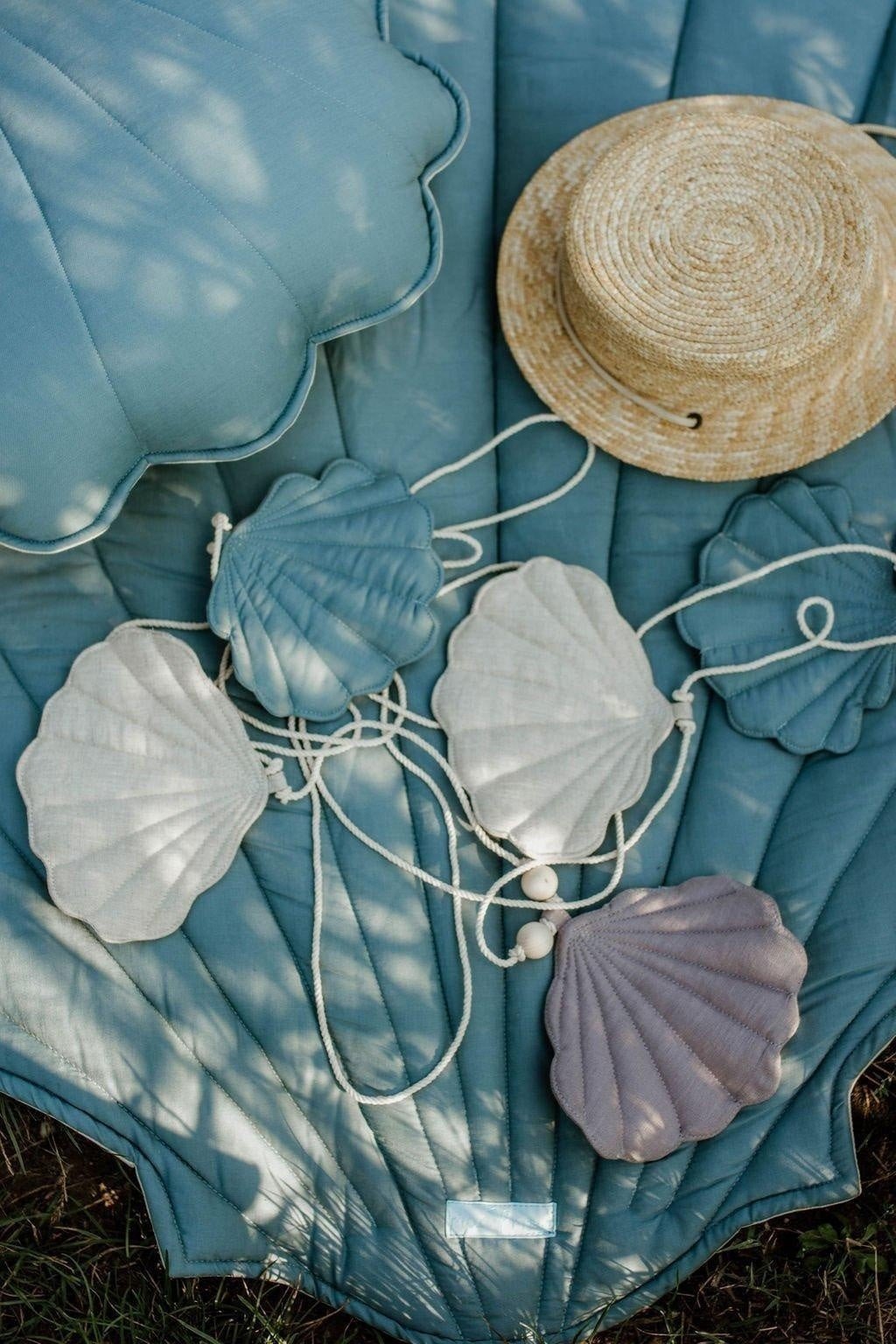3. Shell garland with linen pendants in sky blue, grey, and desert sand on a blue quilt with a straw hat outdoors
