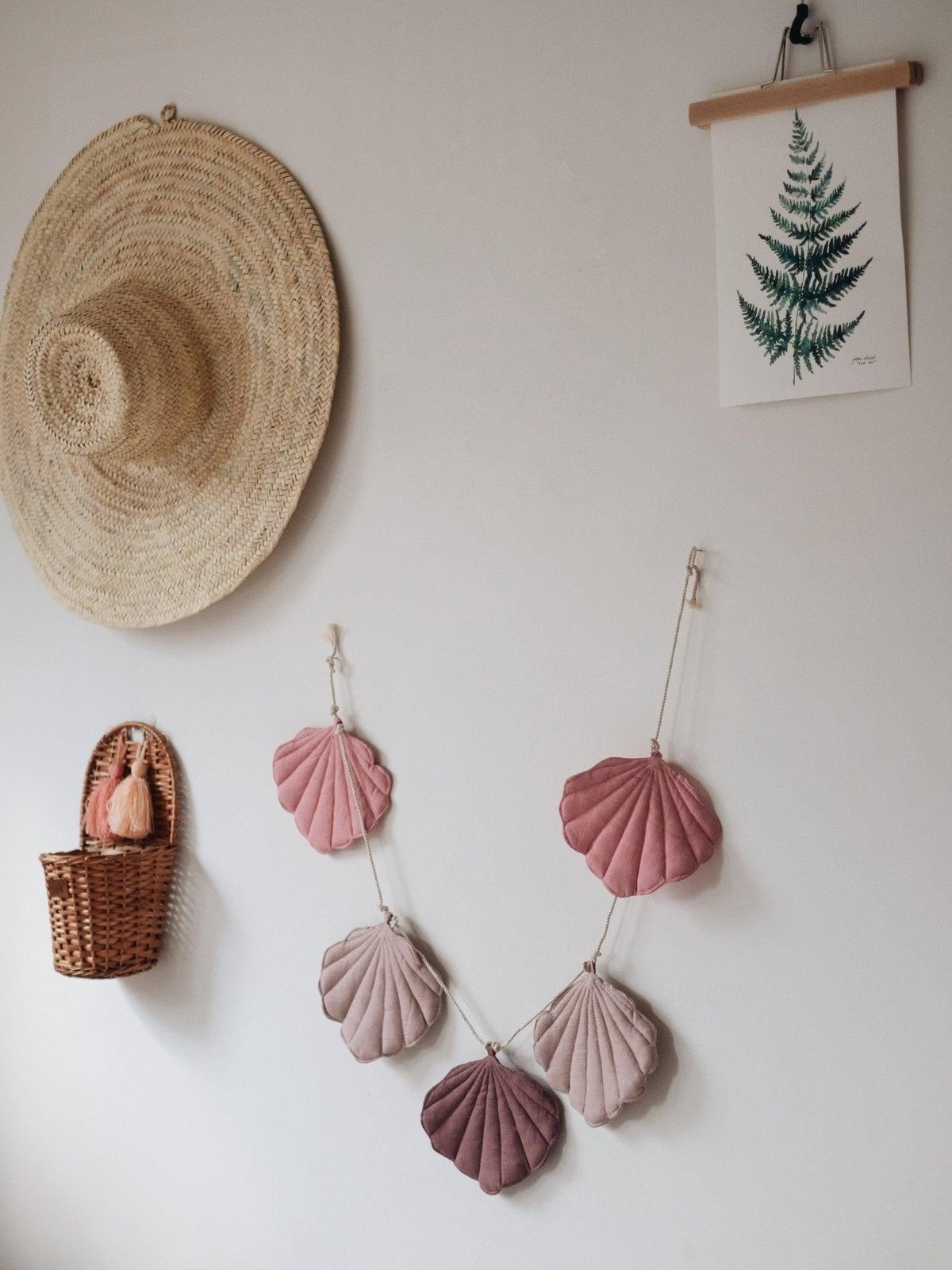 5. Powder pink shell garland with five linen pendants hanging on a wall with a straw hat and fern print decor