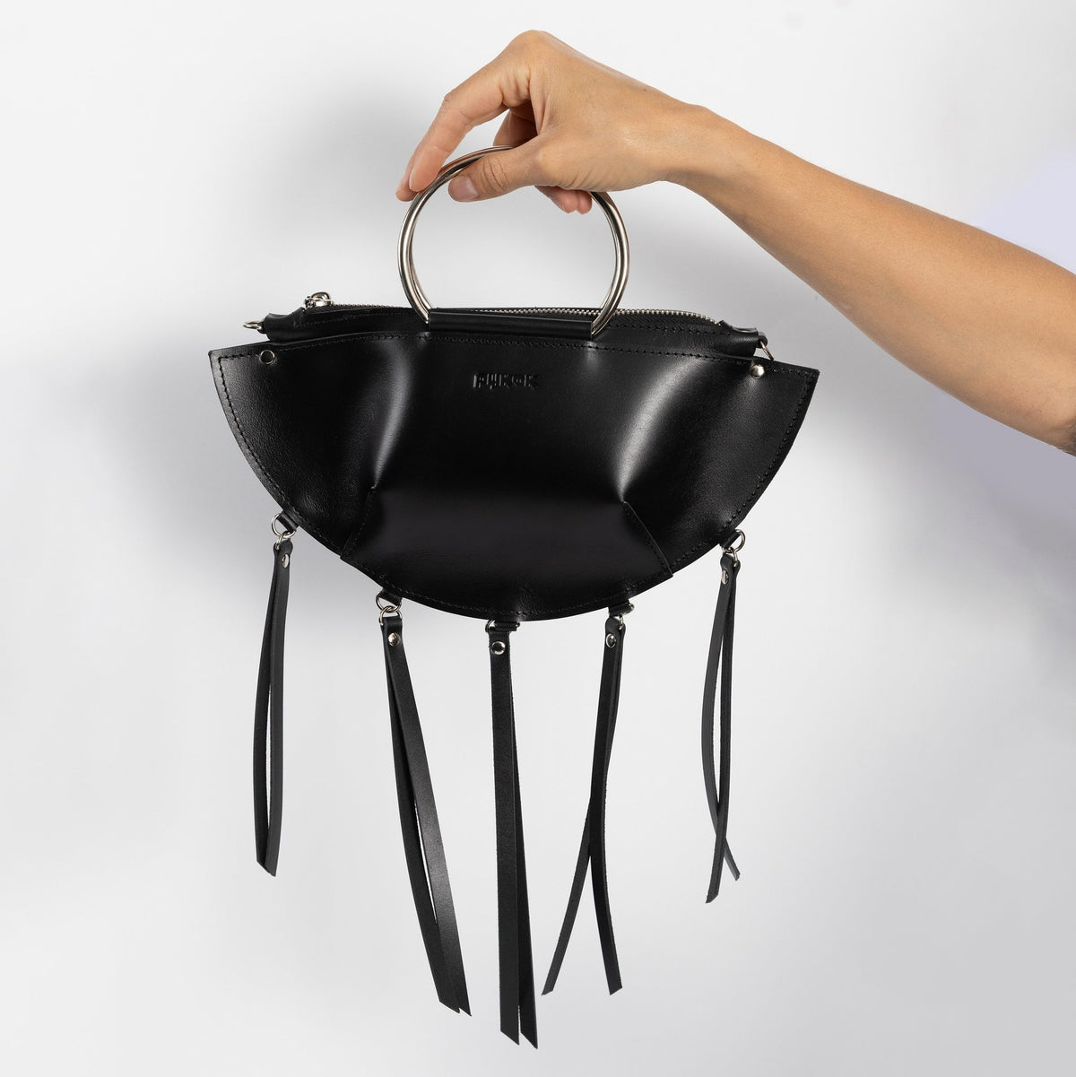 6. Hand holding black leather KAYA handbag with metal ring handles and tassels against a white background
