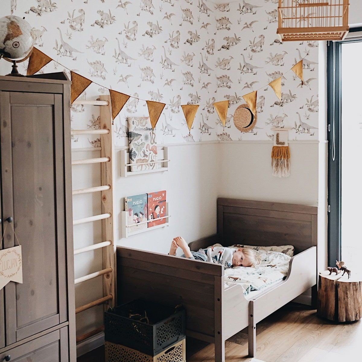 5. Child lying in bed in room with dinosaur wallpaper, wooden furniture, and decorative bunting
