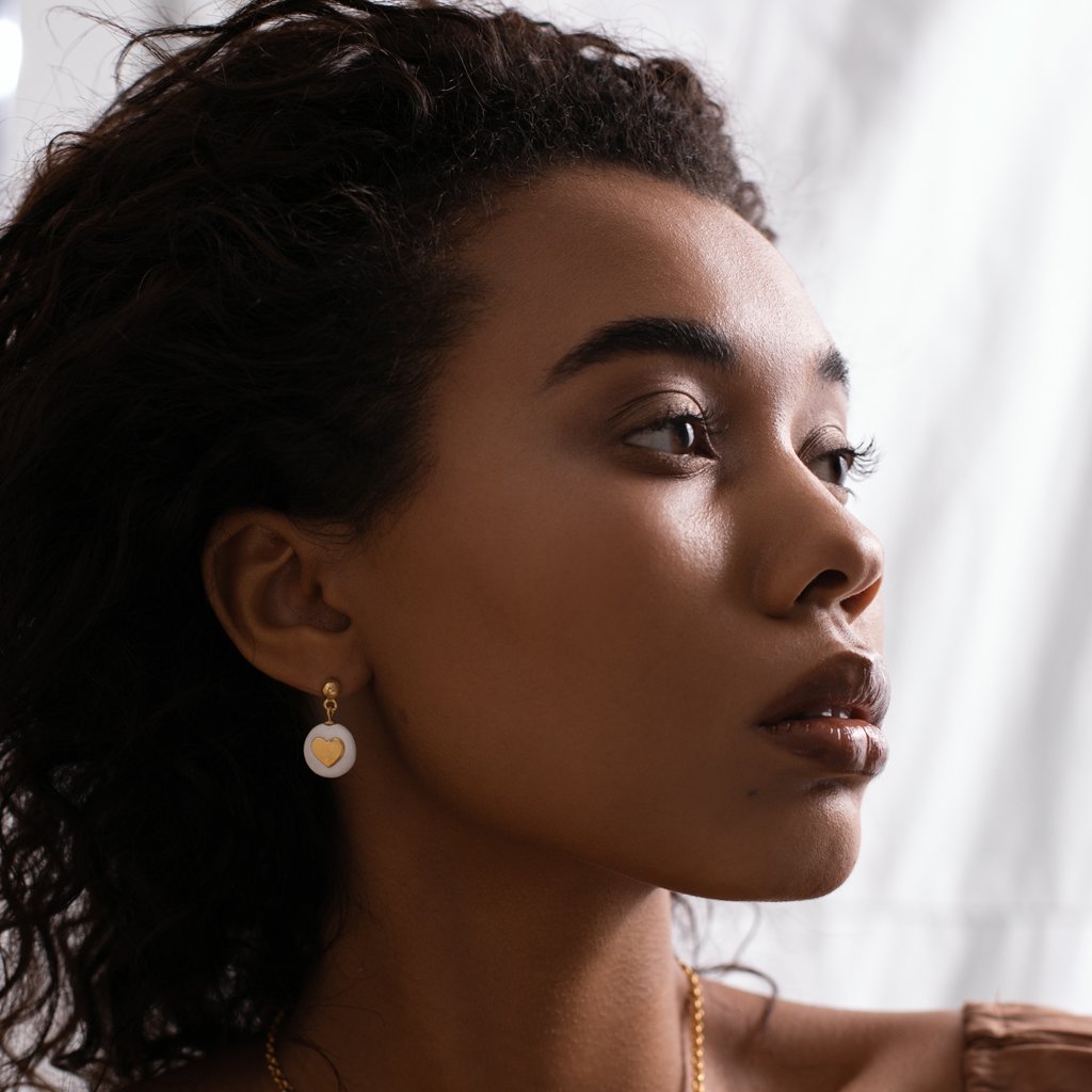 1. Woman wearing SCULP Pure Love Earrings with white ceramic and gold heart detail, shown in lifestyle setting