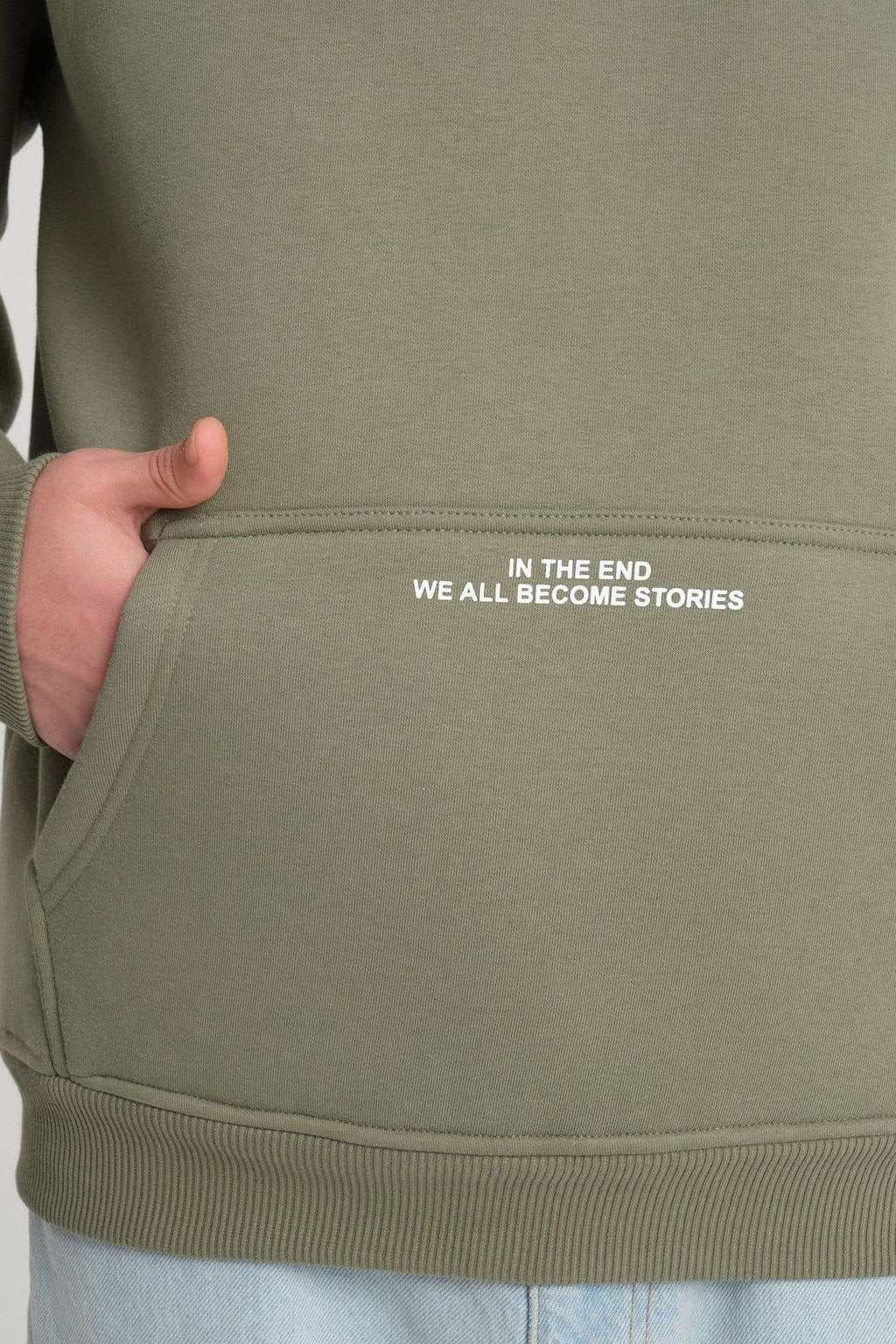 3. Close-up of olive green hoodie pocket with white text 'IN THE END WE ALL BECOME STORIES'