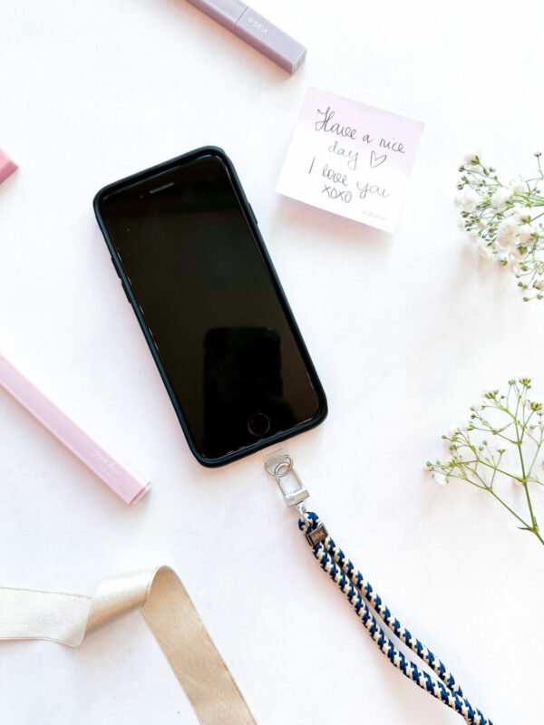 4. Flat lay of SEIK Universal Phone Strap in Sea Edge attached to black phone case with note and flowers