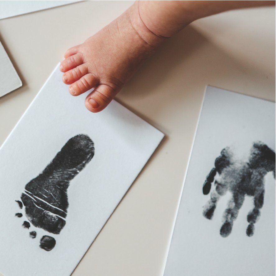 1. Baby's foot next to ink-free footprint and handprint on white paper, showcasing Beebila keepsake print kit