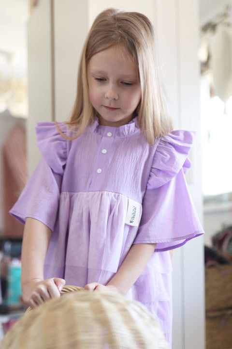 3. Girl in lilac Zezuzulla dress with ruffled sleeves and front snap buttons, standing indoors