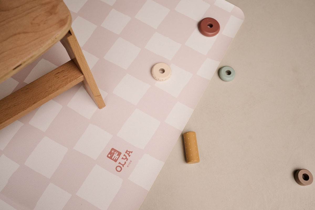 3. Detail of OLYA KIDS logo on pink and beige checkered floor mat with wooden chair leg and toys