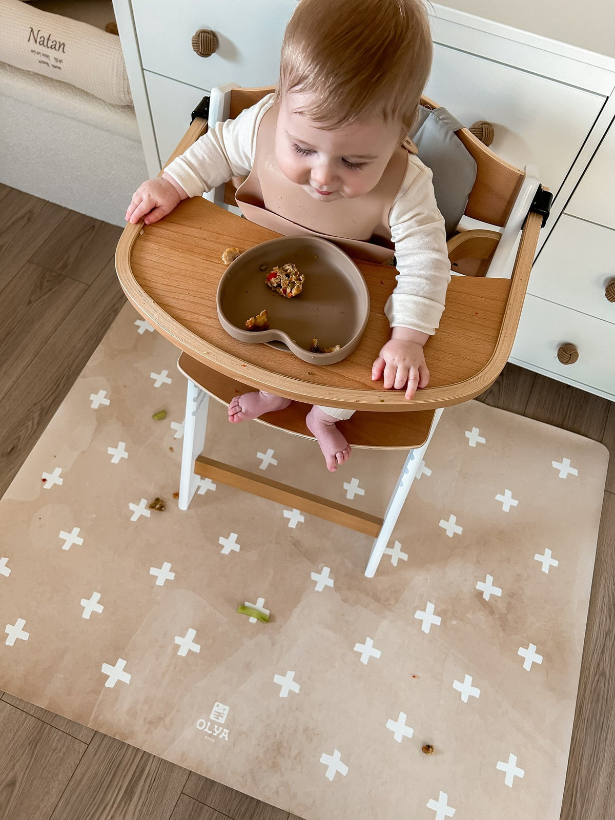1. Baby sitting in high chair on beige OLYA KIDS floor mat with cross pattern, eating from bowl, in cozy home setting