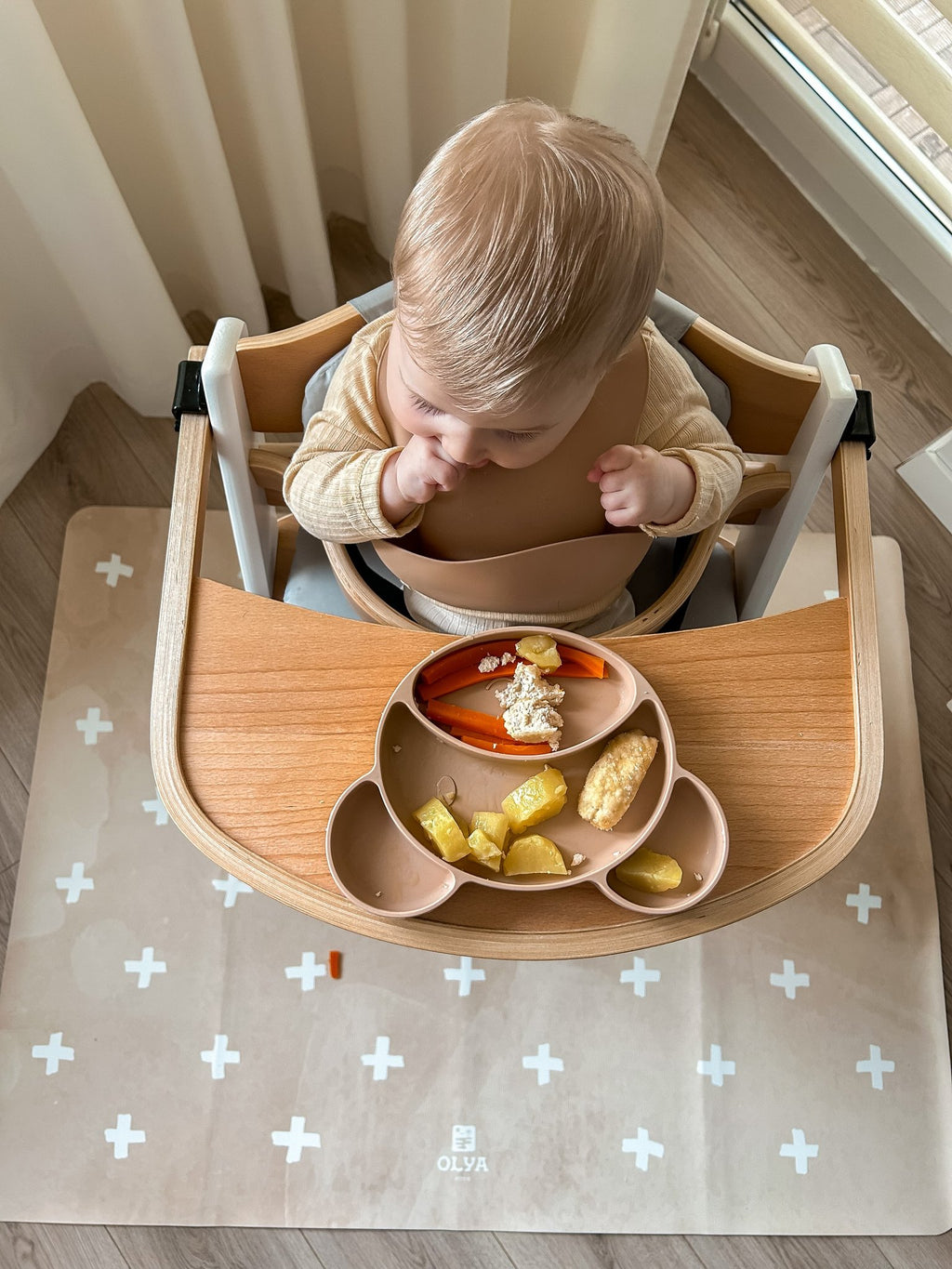 2. Baby in high chair on beige OLYA KIDS floor mat with cross pattern, eating from divided plate, near window