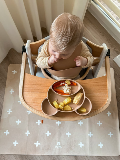2. Baby in high chair on beige OLYA KIDS floor mat with cross pattern, eating from divided plate, near window