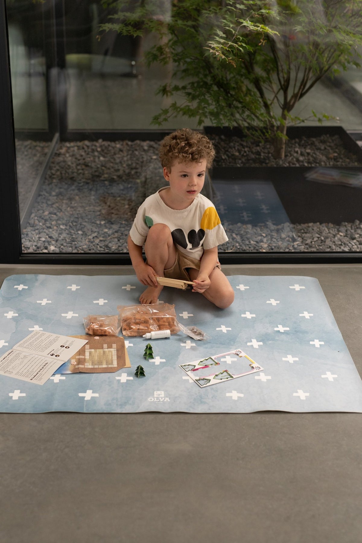 9. Child sitting on blue OLYA KIDS floor mat with white cross pattern, holding craft materials, indoors