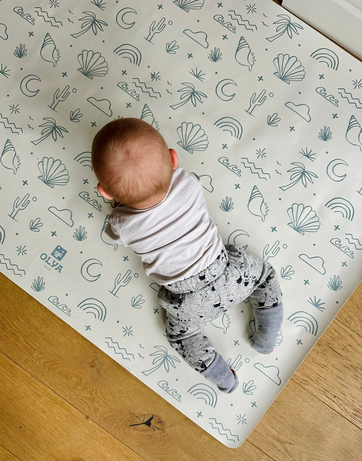 1. Baby lying on OLYA KIDS Palm Beach Azure floor mat with playful patterns, on wooden floor