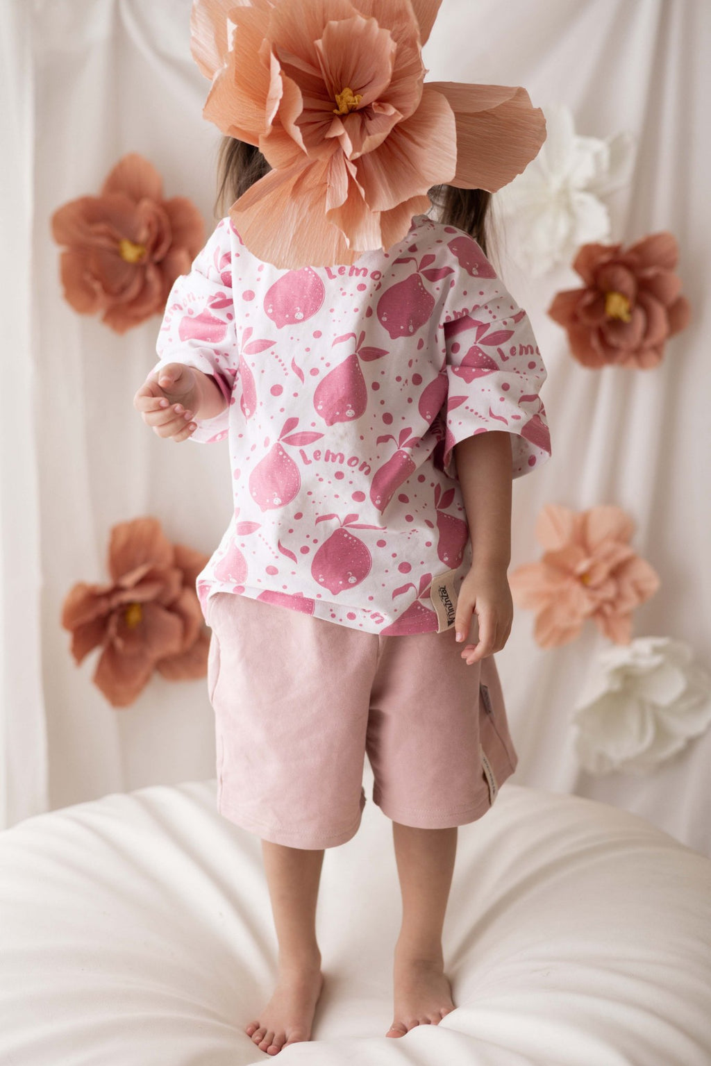 4. Child wearing pastel pink shorts and white printed shirt with large paper flower in front of face against floral backdrop
