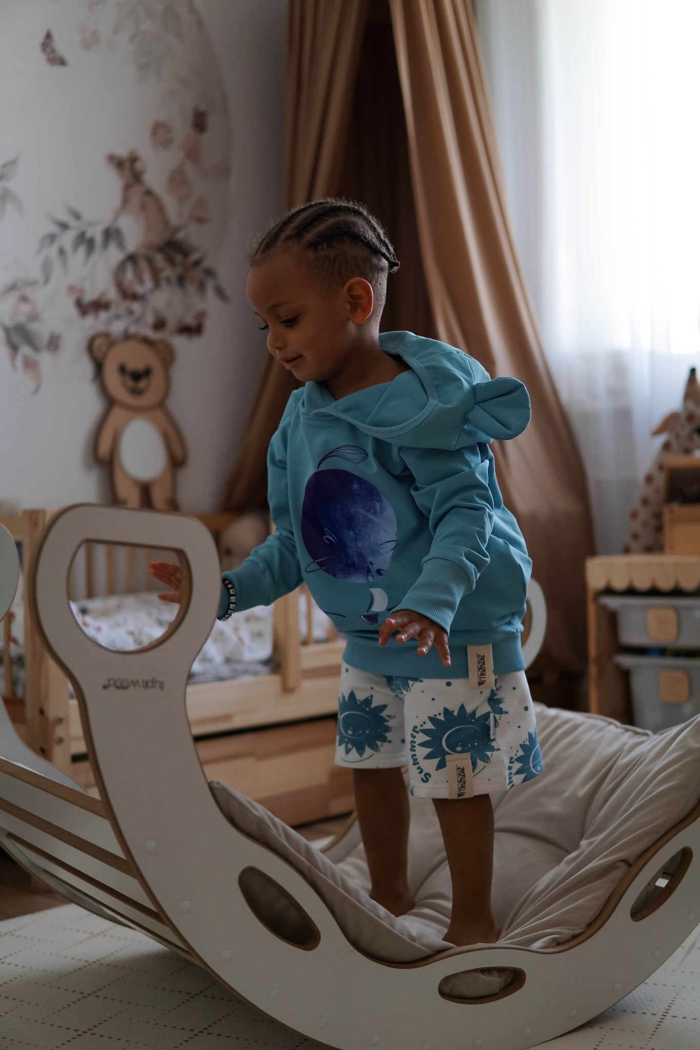 4. Child wearing white shorts with blue sun print and blue hoodie standing on indoor rocker