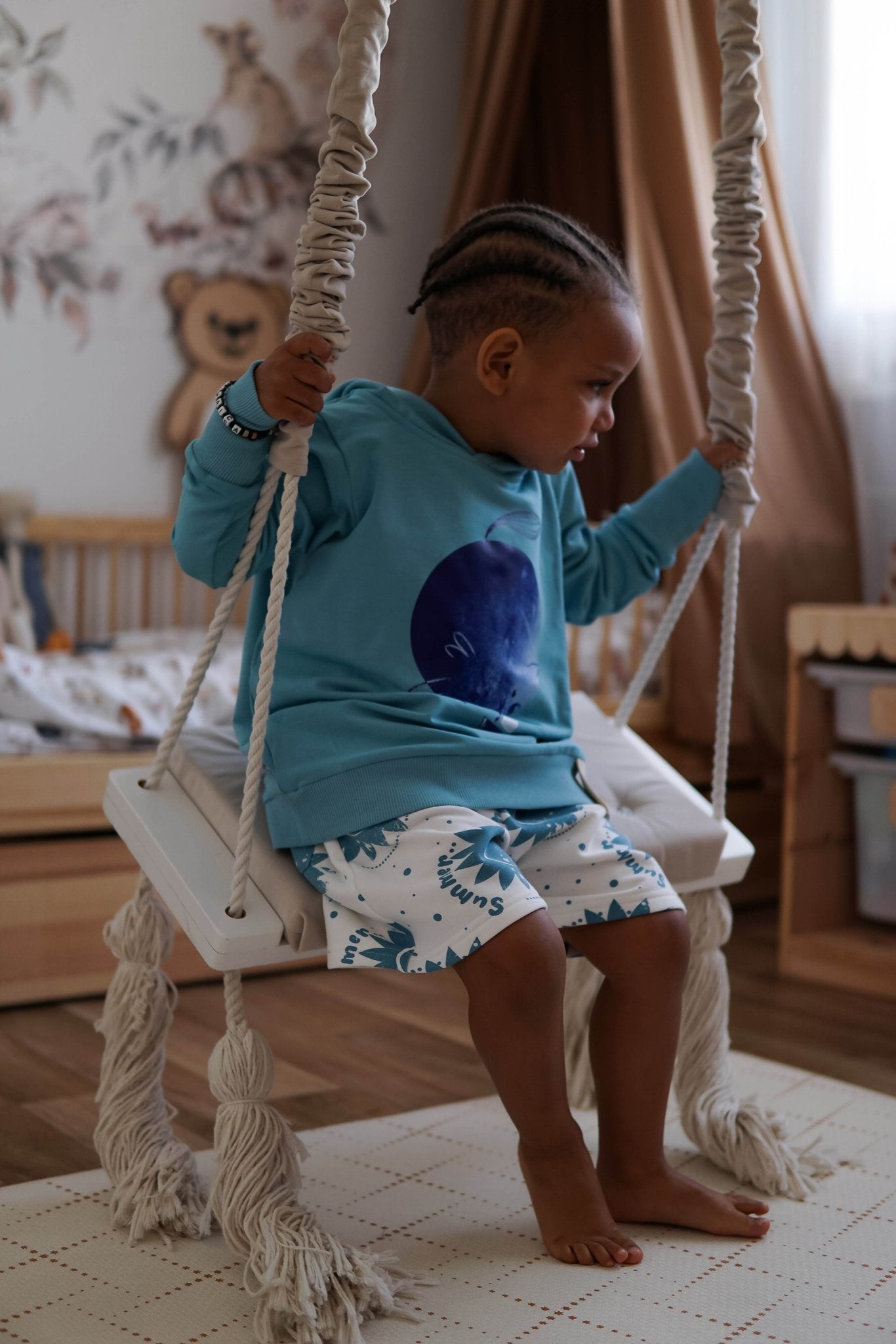 1. Child wearing white shorts with blue sun print and blue hoodie sitting on indoor swing