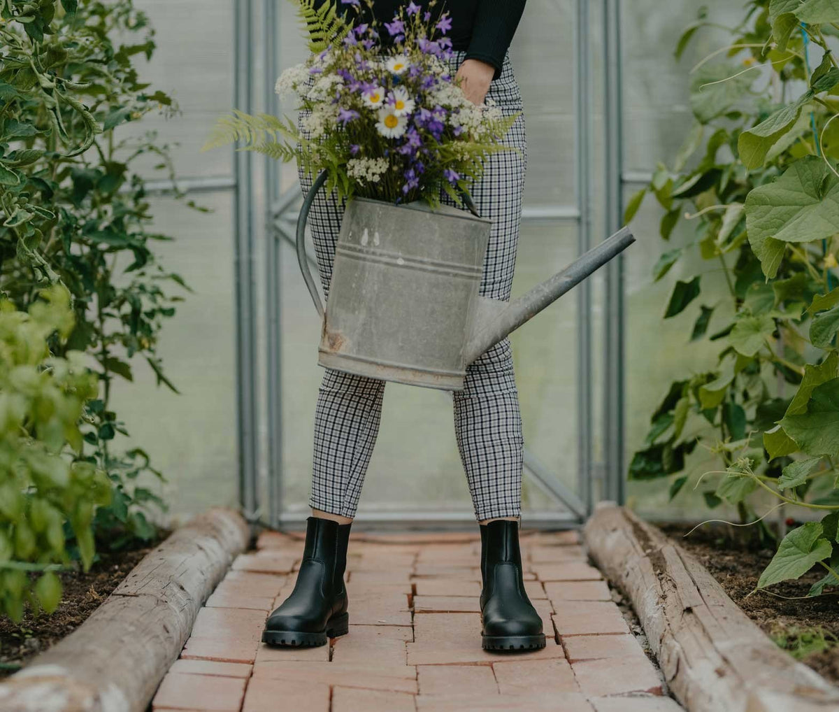 1. Woman wearing KIRA 3.0 Chelsea Boots in black, holding a watering can in a garden setting
