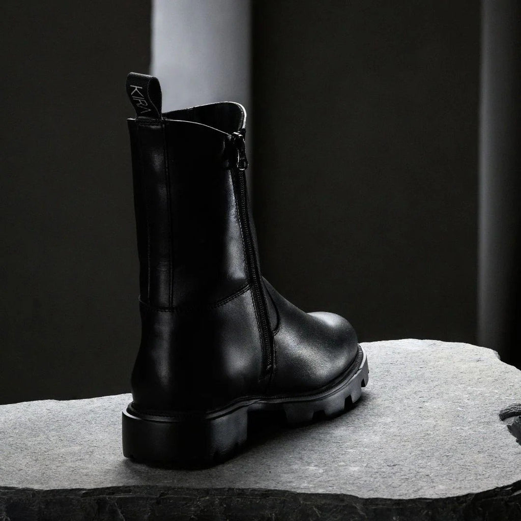 3. Back view of black KIRA Chelsea Rebel boot with smooth zipper and logo pull tab on stone surface