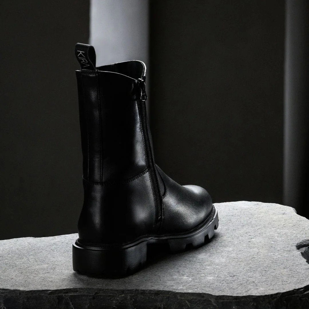 3. Back view of black KIRA Chelsea Rebel boot with smooth zipper and logo pull tab on stone surface