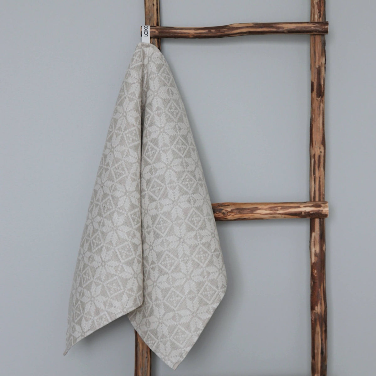 1. Beige kitchen towel with Hiiumaa folk art pattern hanging on rustic wooden ladder, made of 52% linen and 48% cotton by LOKO, designed in Estonia