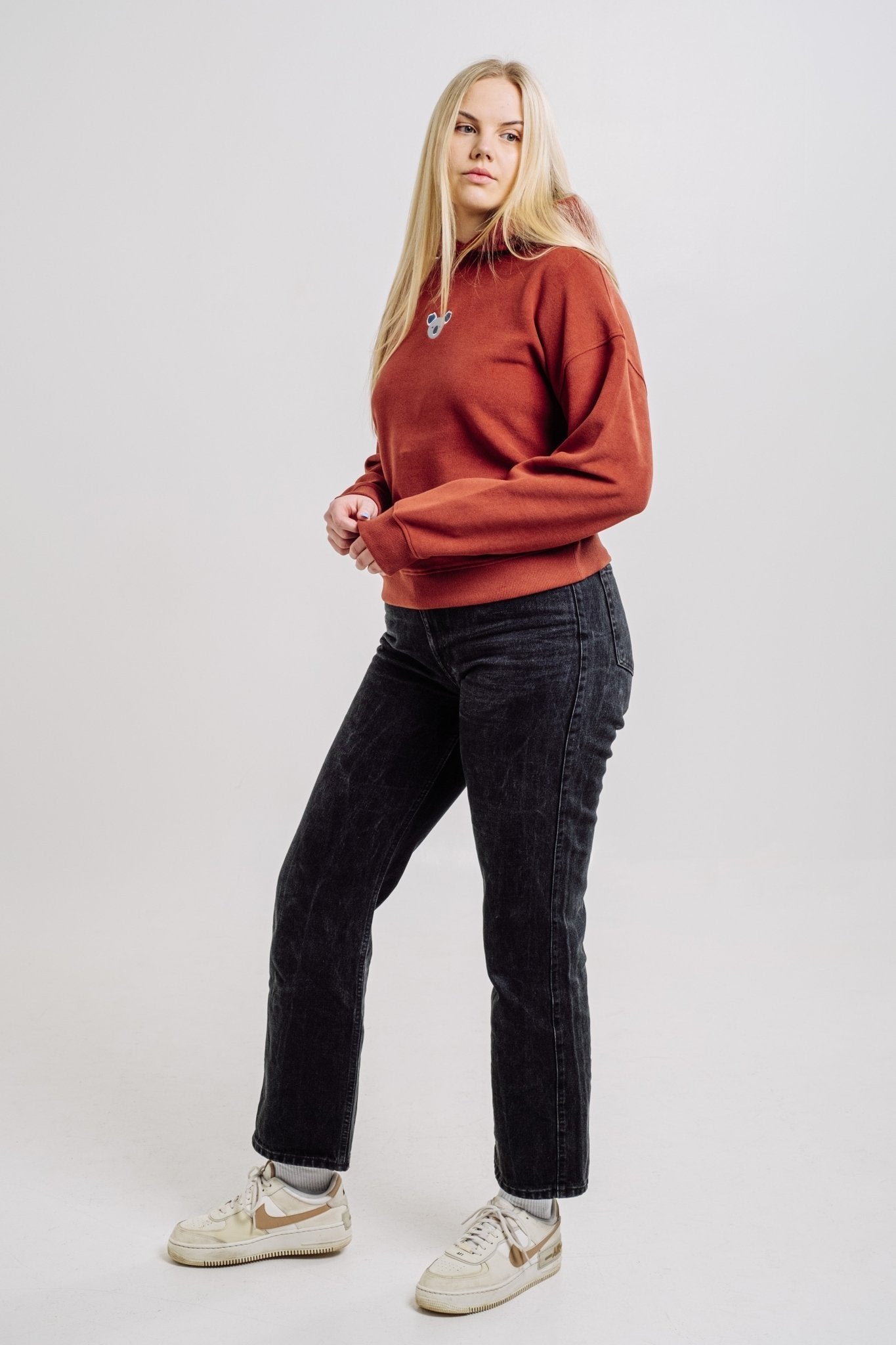 1. Woman wearing Sossu Koala Hoodie in terracotta, styled with black jeans and sneakers, studio setting